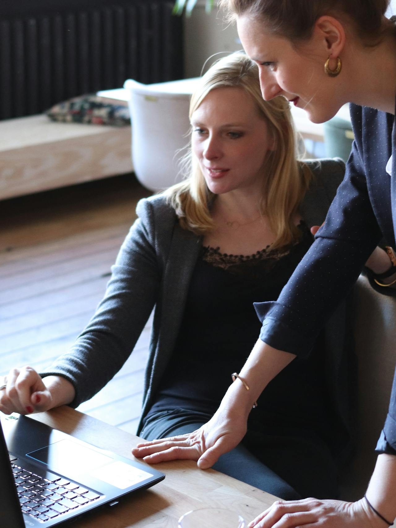 Two professional women discuss a project using a laptop in a modern office environment.